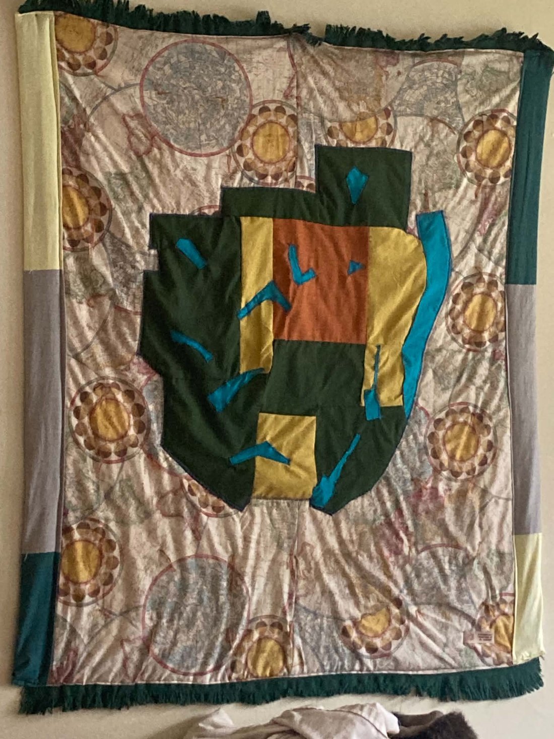 Appliqué quilt of the Adirondack Park in green, gold, rust, and teal fabric on a vintage map print backing with dark green fringe